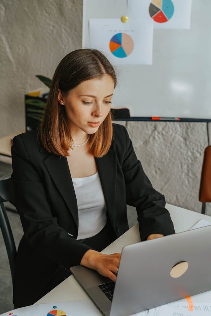 Confident businesswoman working on a laptop in a modern office environment with charts in the background.